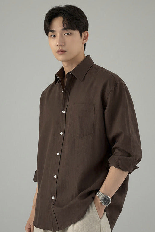A man wearing a dark brown button-up shirt and beige trousers stands in a modern kitchen, holding a large brown leather tote bag.