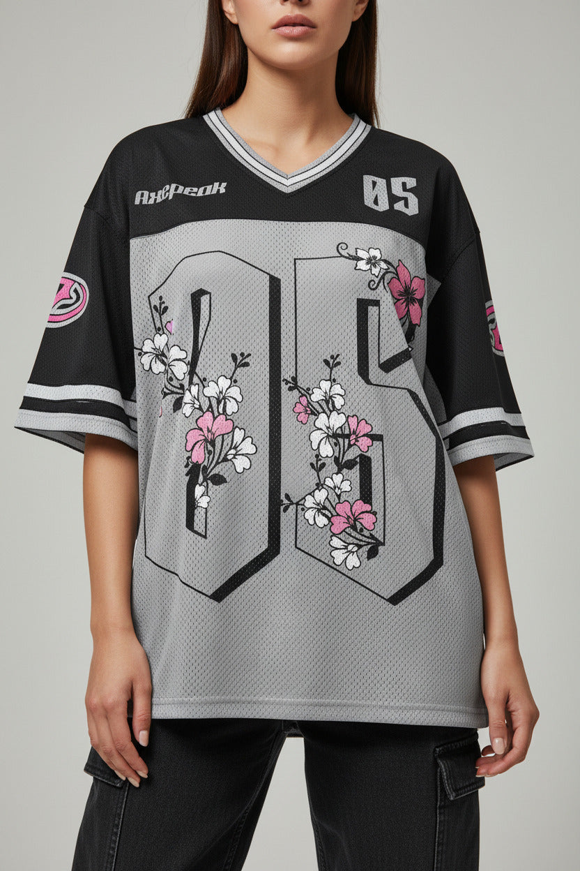 A short-sleeve athletic jersey laid flat on a textured gray concrete surface. The jersey has a black upper chest and shoulders, a gray mesh lower body, and pink and white cherry blossom floral graphics over a large black number 85 on the front.