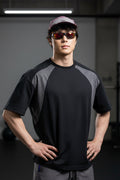 A man wearing a black and gray short-sleeve athletic t-shirt, gray shorts, a pink cap, and orange-tinted sunglasses, standing outdoors against a large rock.