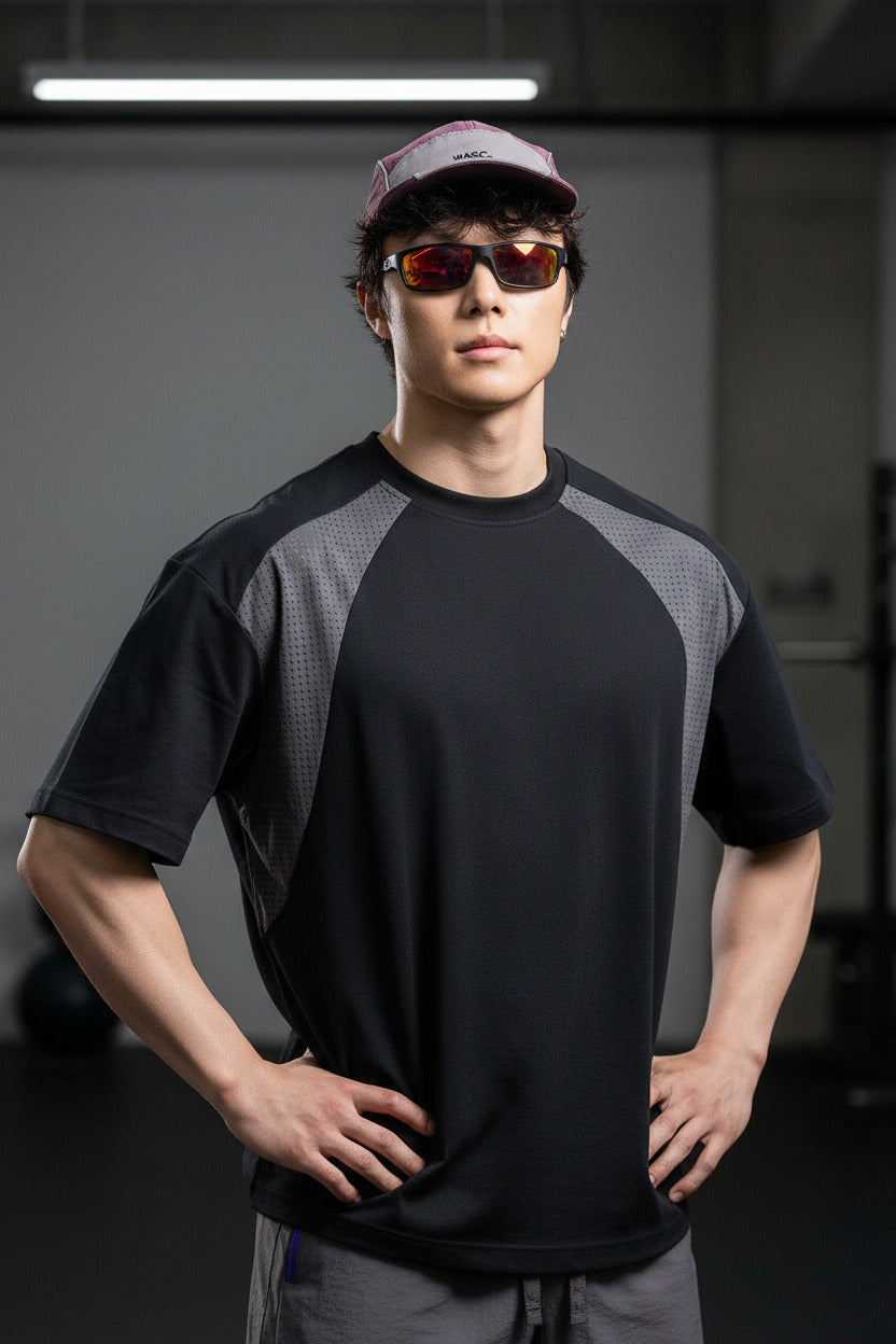 A man wearing a black and gray short-sleeve athletic t-shirt, gray shorts, a pink cap, and orange-tinted sunglasses, standing outdoors against a large rock.