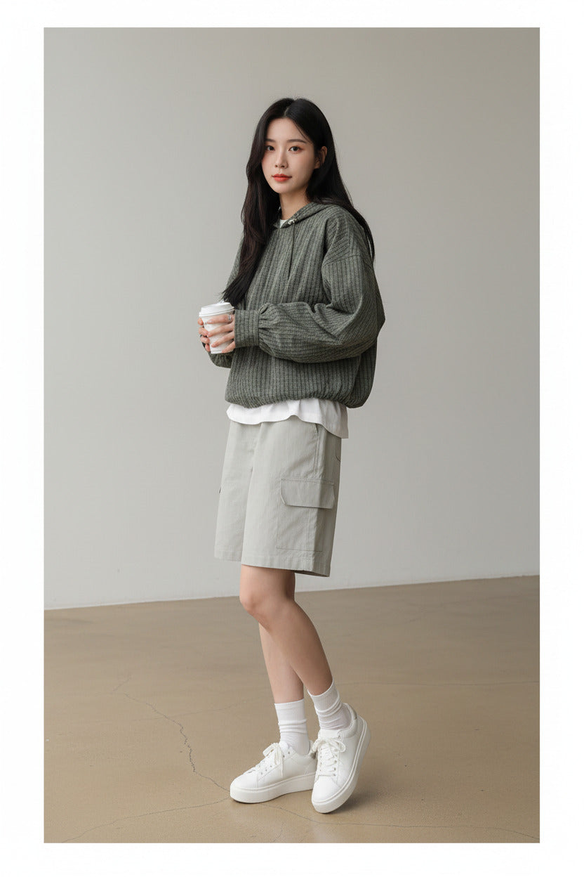 A woman wearing an oversized green and black checkered zip-up hoodie with long sleeves, paired with light gray shorts, holding a coffee cup in a cafe setting.
