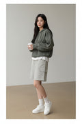 A woman wearing an oversized green and black checkered zip-up hoodie with long sleeves, paired with light gray shorts, holding a coffee cup in a cafe setting.