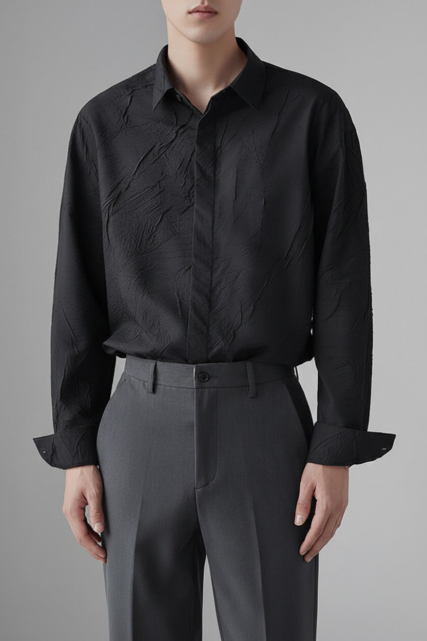A man wearing a black long-sleeve button-down shirt with a unique crinkled or textured fabric finish, paired with dark gray trousers, shown from the neck down against a neutral gray background.