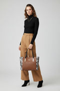 A structured tote bag with a brown crocodile-embossed center panel, black handles, and a snakeskin-patterned side panel, displayed on a black book in front of a framed fashion poster.