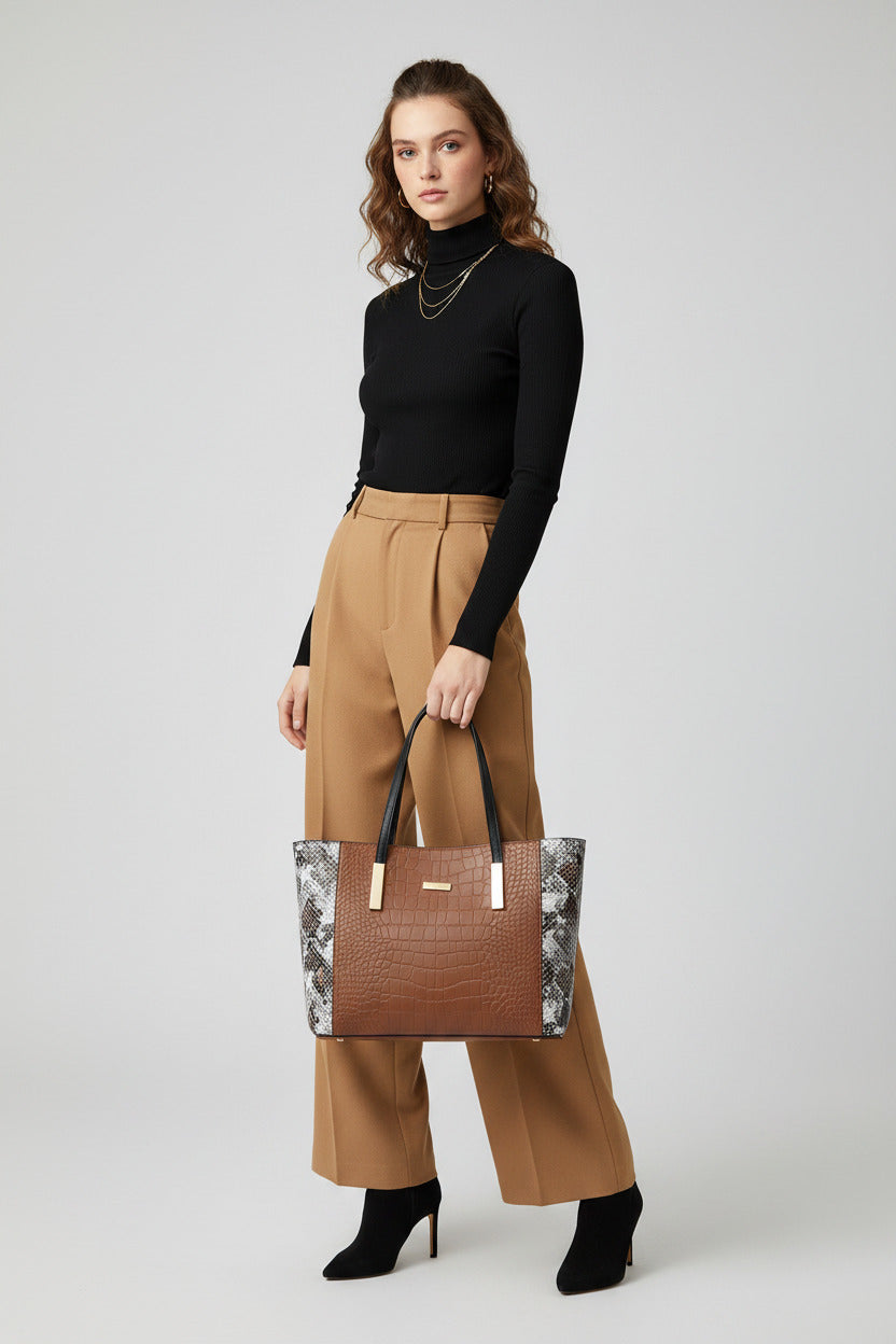A structured tote bag with a brown crocodile-embossed center panel, black handles, and a snakeskin-patterned side panel, displayed on a black book in front of a framed fashion poster.