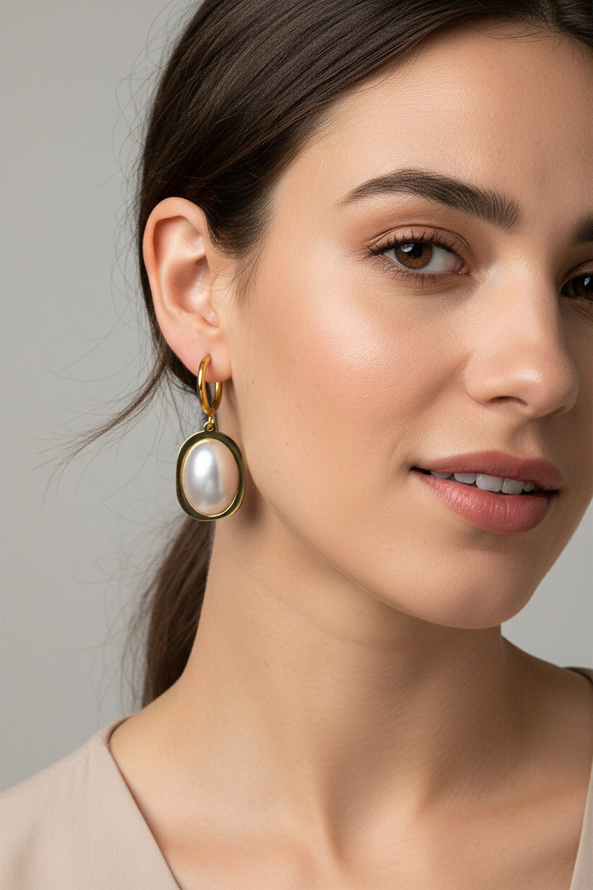 A pair of gold hoop earrings with oval gold frames and large white pearl-like drops, displayed on a soft beige fabric background.