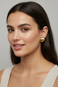 Two small gold stud earrings with organic, rounded shapes and textured finishes, displayed on a beige ceramic plate next to a white card.