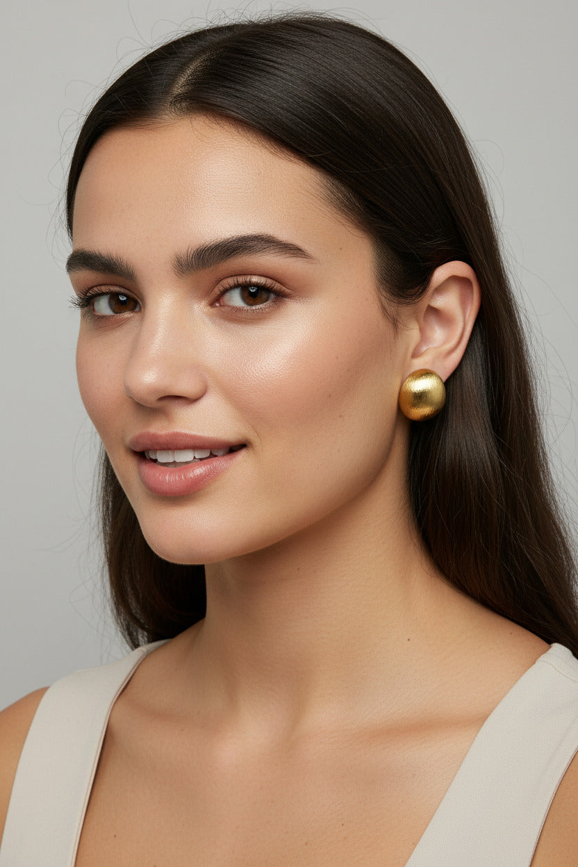 Two small gold stud earrings with organic, rounded shapes and textured finishes, displayed on a beige ceramic plate next to a white card.