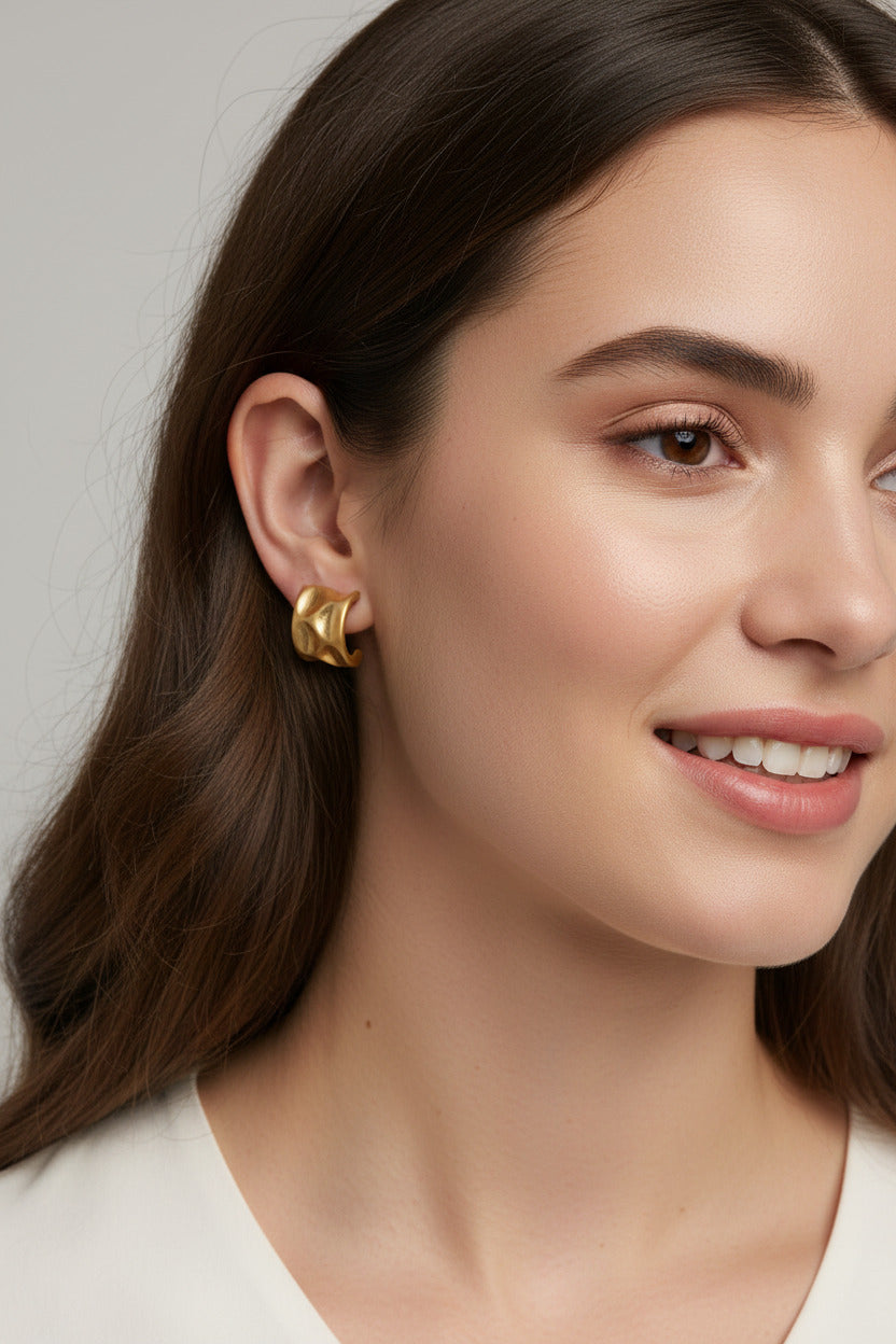 A pair of textured gold stud earrings displayed on a small wooden block, placed on top of an open book with blue denim visible in the corner.