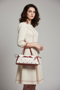 A close-up of a handbag with a white textured body, small red bow and dot pattern, glossy red handles and trim, and a dangling heart-shaped charm engraved with 'Spectro Homo London'.