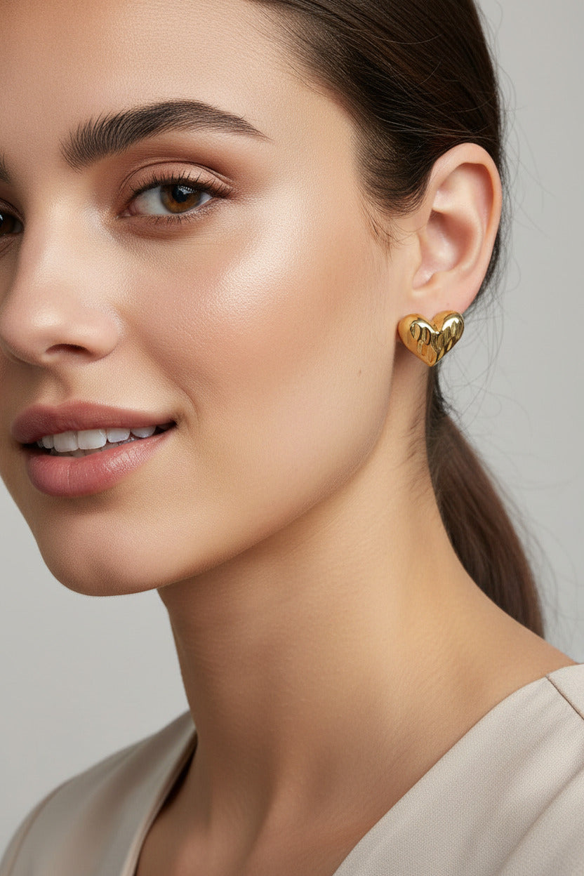 A pair of gold heart-shaped stud earrings with a textured, ridged surface, resting on a draped beige fabric background.