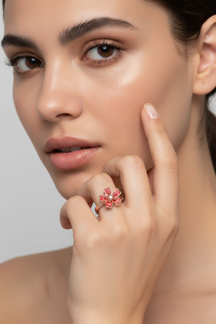 A rose gold ring featuring a cherry blossom flower with red enamel petals and a clear central gemstone, shown on a soft pink background with a small '360-degree free rotation' text overlay.