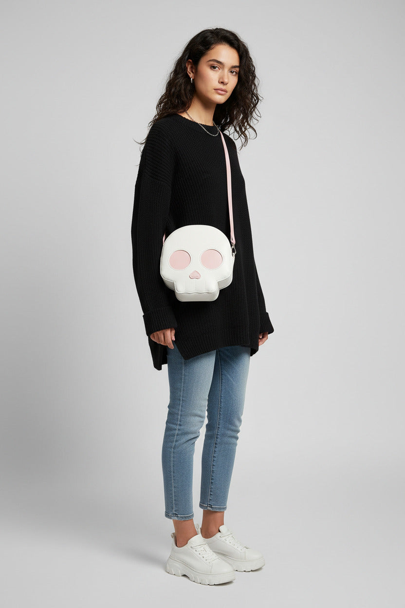 A person wearing a dark navy knit sweater carries a small skull-shaped crossbody bag. The bag is off-white with pink circular eye cutouts, a pink nose, and a light pink strap.
