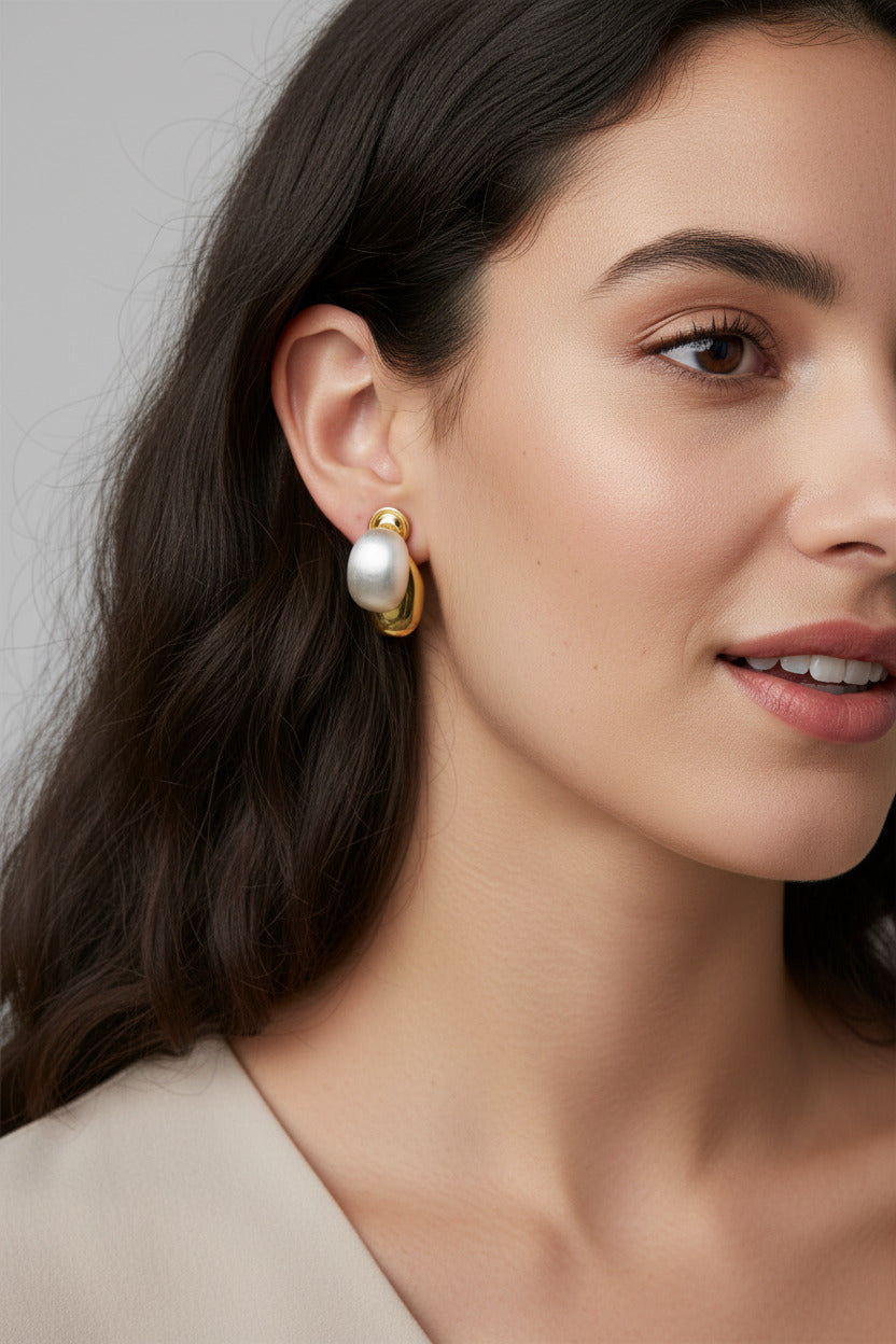 A pair of gold-toned stud earrings with oval milky white stones, displayed on a brown ribbed knit surface next to a white textured fabric and a cream-colored book corner.