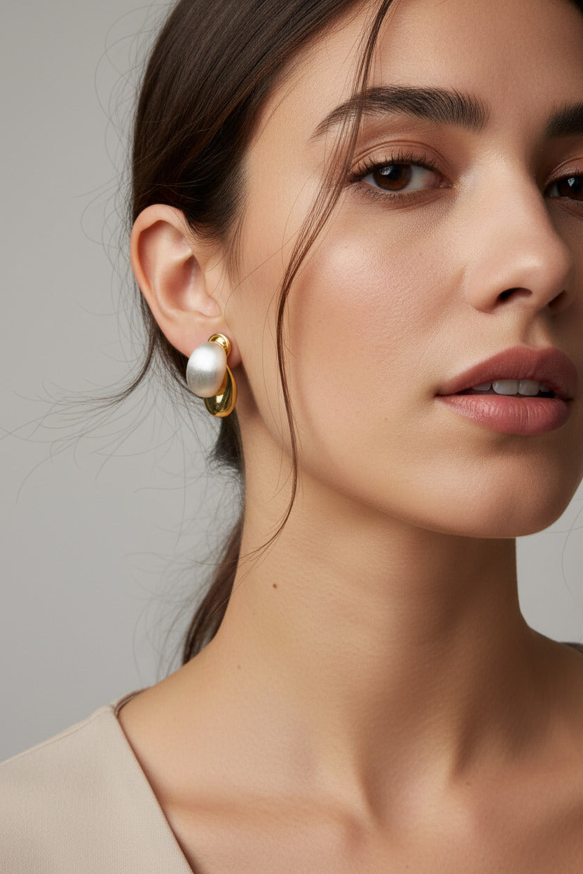 A pair of gold-toned stud earrings with oval milky white stones, displayed on a brown ribbed knit surface next to a white textured fabric and a cream-colored book corner.