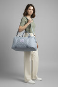 A woman wearing a beige baseball cap and a light yellow-green t-shirt is shown from behind, carrying a light blue-gray quilted duffel bag with a gray strap. The setting is a bright room with a window, blinds, and a black sideboard.