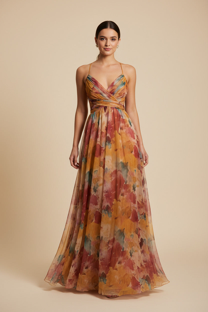 A woman takes a mirror selfie outdoors at night wearing a flowing sleeveless maxi dress with a multicolor watercolor floral pattern in orange, yellow, pink, and blue. She is standing on a lawn with lit dinner tables and palm trees in the background.
