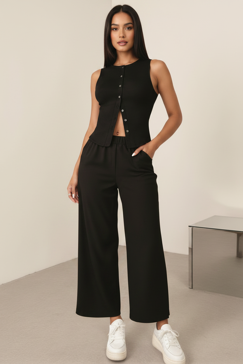 A woman modeling a black sleeveless button-front top and matching black wide-leg pants with white sneakers, standing in a minimalist indoor room with a small table.