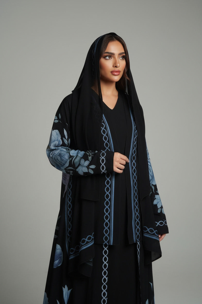 A woman models a long black abaya dress with blue floral prints and chain-link details, standing in a room with a yellow-green velvet sofa and white paneled walls.