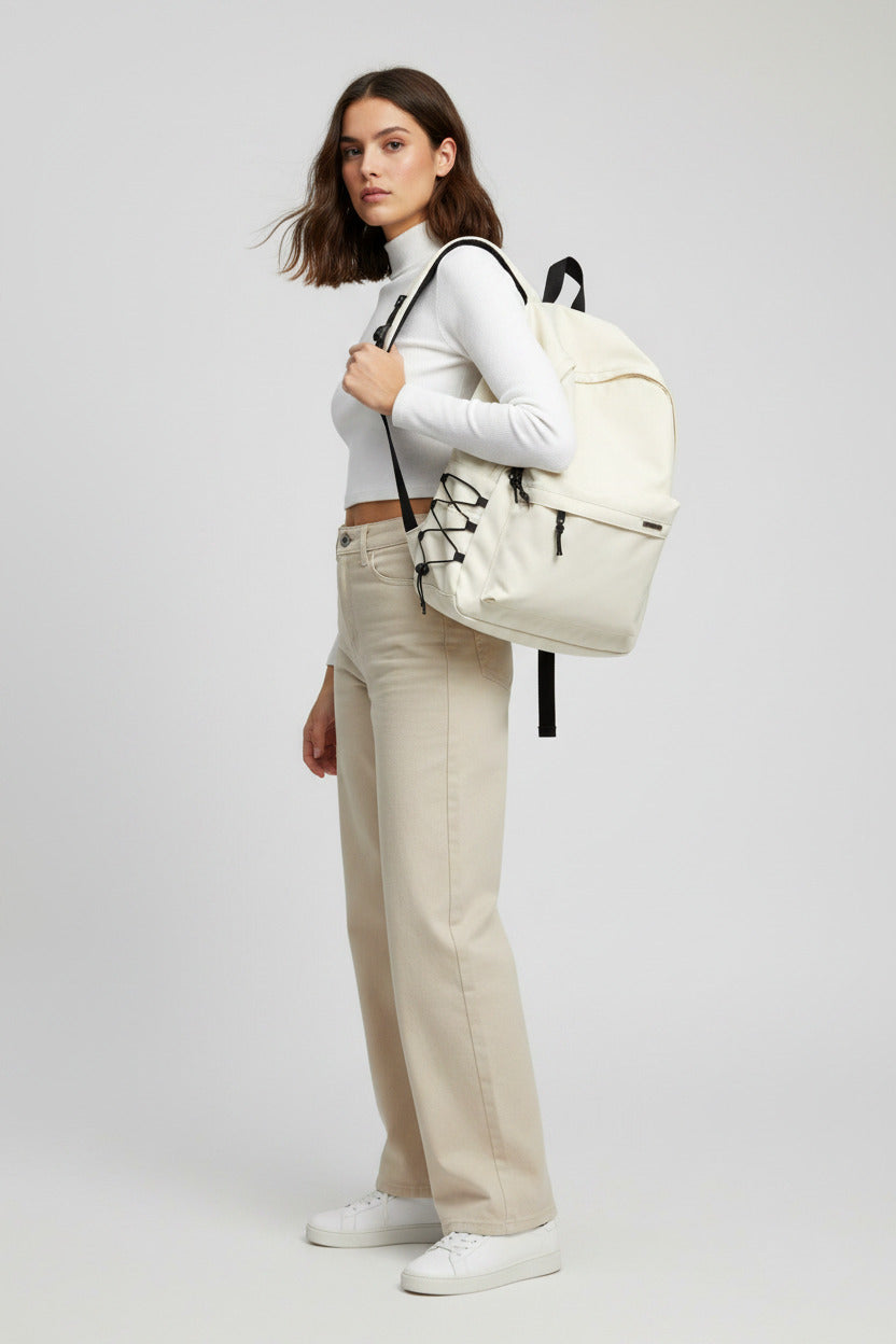 An off-white canvas backpack with black accents, worn by a person wearing a gray sweatshirt and blue jeans, set against a split white and black slatted background.