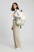 An off-white canvas backpack with black accents, worn by a person wearing a gray sweatshirt and blue jeans, set against a split white and black slatted background.