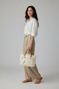 A person wearing a white long-sleeve shirt and beige skirt carries a large cream-colored woven tote bag with brown leather shoulder straps in an indoor setting.
