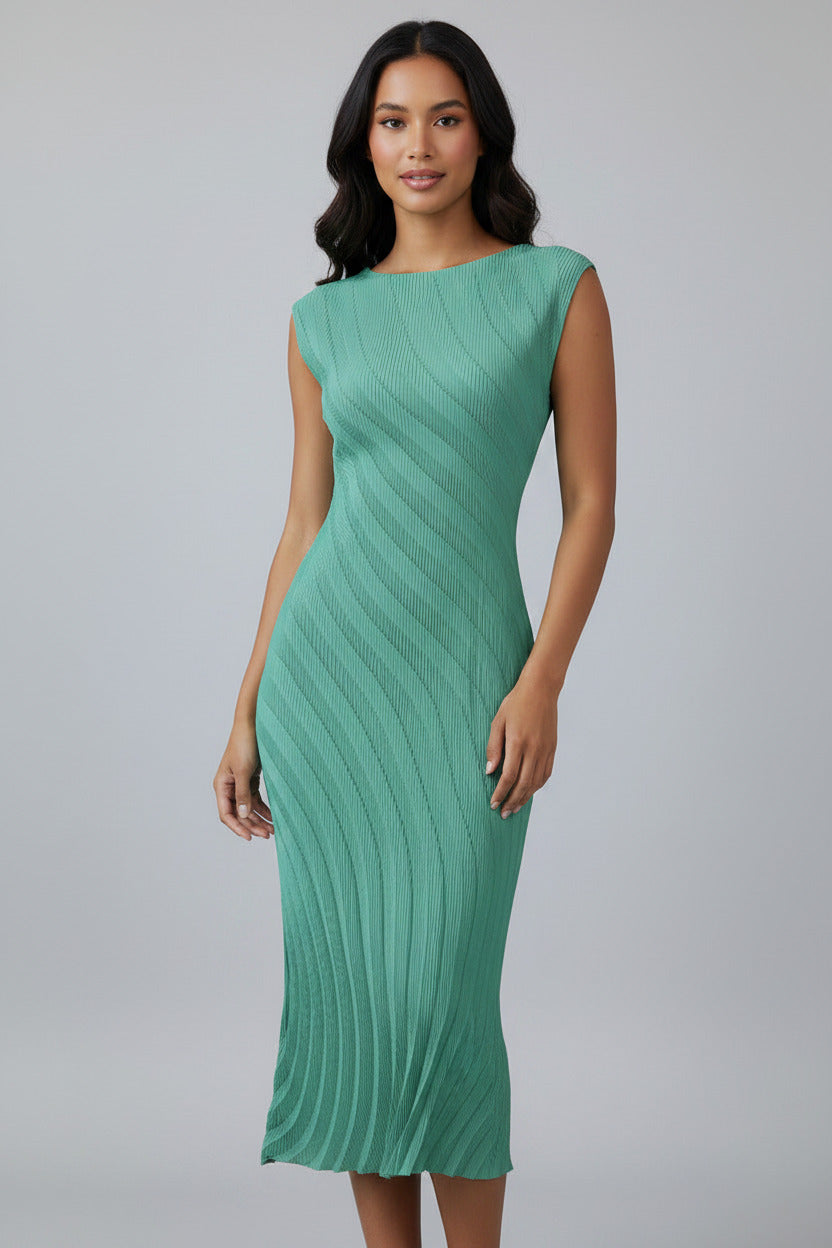 A woman wearing a sleeveless, knee-length green ribbed knit dress, standing in a minimalist room with a white wall, light wood floor, and a small white side table with a vase of branches.
