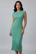 A woman wearing a sleeveless, knee-length green ribbed knit dress, standing in a minimalist room with a white wall, light wood floor, and a small white side table with a vase of branches.
