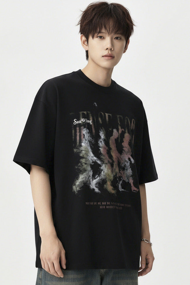 A young man wearing a black graphic t-shirt with a colorful abstract print, blue jeans, and silver headphones, posing against a light gray studio background.