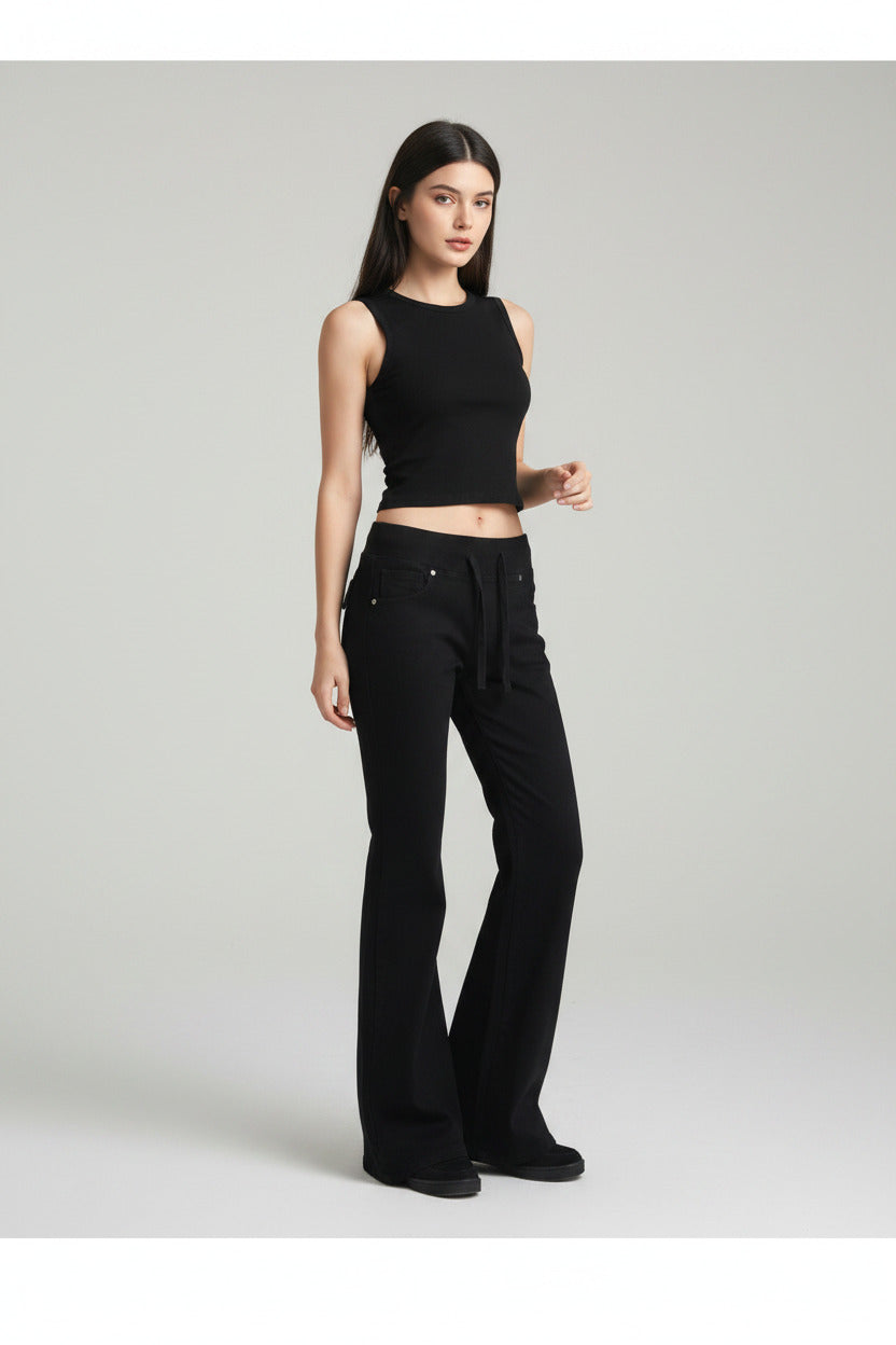 A person wearing black wide-leg flare jeans with a drawstring waist, standing in a room with a wooden floor, desk, and computer monitor in the background.