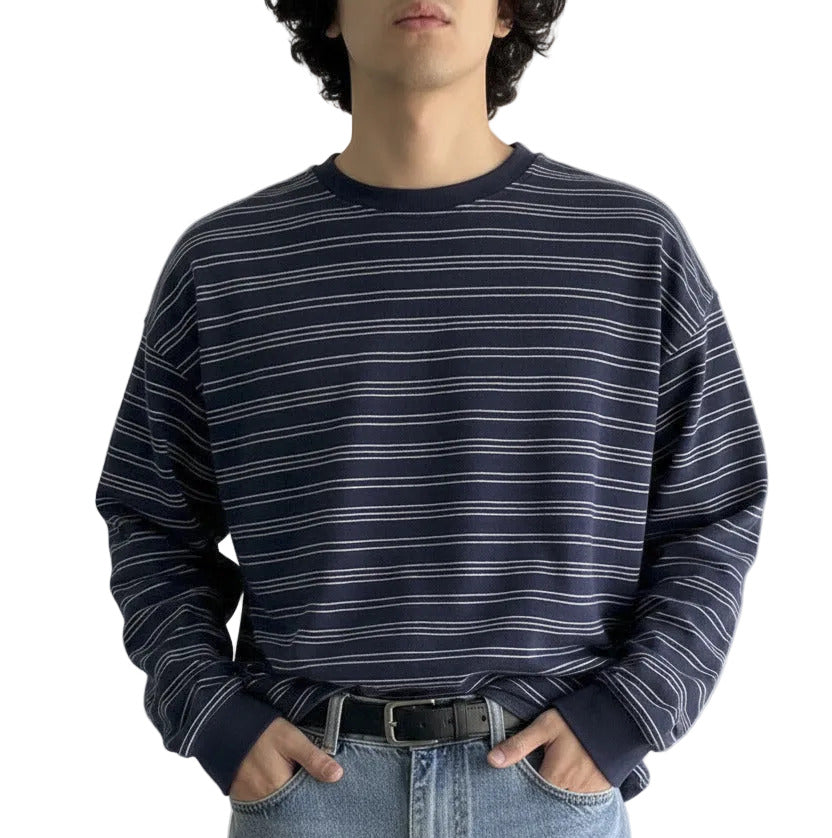 A person wearing a navy blue long-sleeve crewneck shirt with thin white horizontal stripes, paired with light-wash blue jeans, standing against a plain off-white wall.