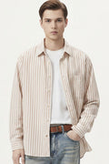 A man wearing a beige long-sleeve button-up shirt with thin vertical brown stripes, layered over a white t-shirt and paired with blue jeans and a brown belt, against a plain off-white background.