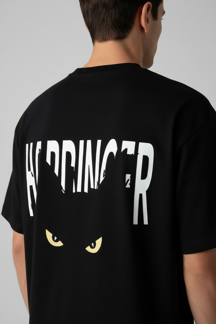 A black short-sleeve t-shirt laid flat on a textured concrete floor, featuring a white graphic of the word 'HARDINGER' with a black cat silhouette and yellow eyes underneath.