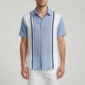 A man wearing a short-sleeve button-up shirt with a light blue and white color-blocked design, featuring dark blue vertical stripes down the front, set against a blurred beach background with palm leaves.