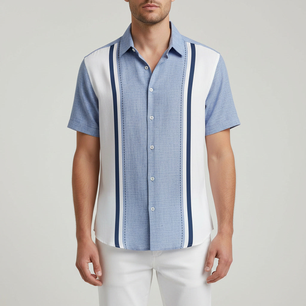 A man wearing a short-sleeve button-up shirt with a light blue and white color-blocked design, featuring dark blue vertical stripes down the front, set against a blurred beach background with palm leaves.