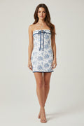 A white strapless mini dress with a blue floral toile pattern and a matching blue tie-front bow, displayed on a clear mannequin against a white background.