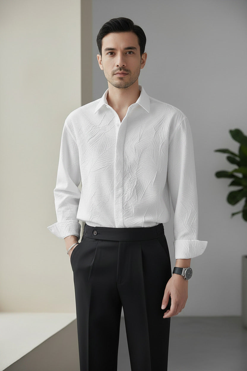 A man wearing a white long-sleeve button-up shirt with a subtle textured pattern, paired with black trousers, standing against a light gray background.