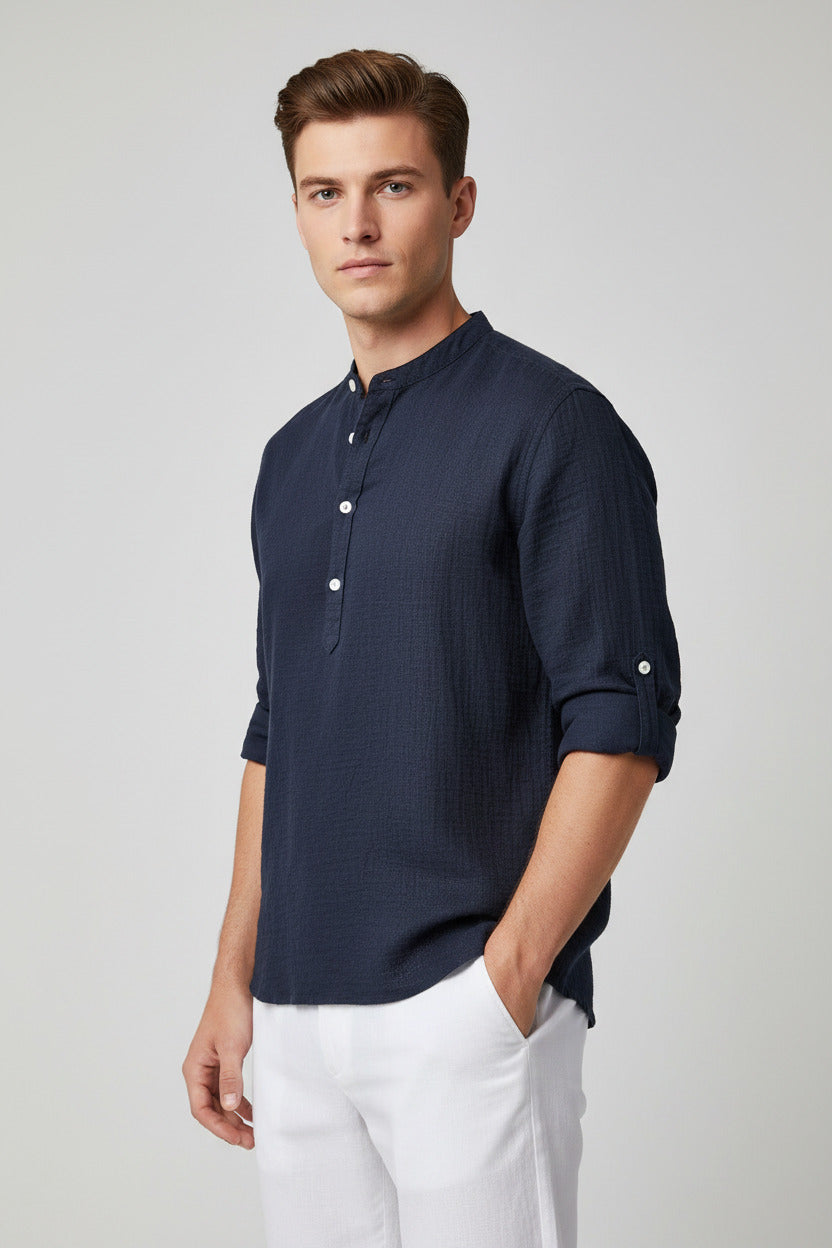 A man wearing a dark navy blue textured short-sleeve henley shirt with rolled-up sleeves and white pants, holding a straw hat against a plain white background.