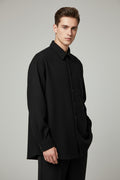 A black long-sleeve button-up shirt with a pointed collar and chest pocket, laid flat on a black mesh office chair over a patterned floor.